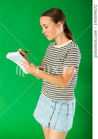 Teen elegant girl dressed in summer T-shirt and blue jeans skirt posing against green backdrop holding documents or writing notes in notebook Teen elegant girl dressed in summer T-shirt and blue jeans skirt posing against green backdrop holding documents or writing notes in notebook 140894872