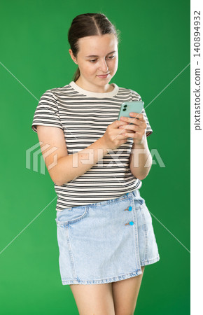 Teenage girl in denim skirt looks at smartphone screen green screen background. Chroma key compositing Teenage girl in denim skirt looks at smartphone screen green screen background. Chroma key compositing 140894932