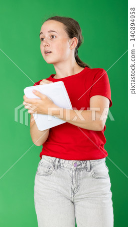 Teen young girl dressed in summer red T-shirt and blue jeans posing against green backdrop holding documents or writing notes in notebook 140894958