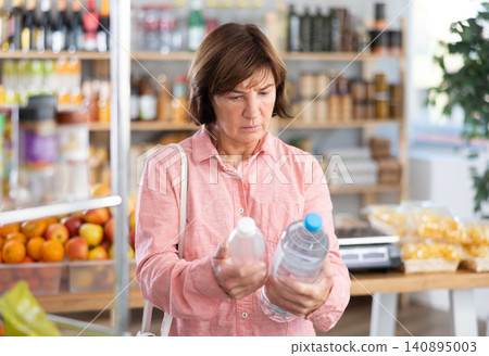 Female buyer carefully reads labels on water bottles - she chooses the best product in supermarket 140895003