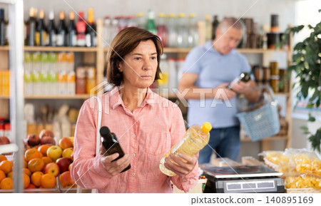 Mature woman buyer looks thoughtfully at bottles of vegetable oil, man client in background Mature woman buyer looks thoughtfully at bottles of vegetable oil, man client in background 140895169