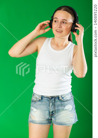 European teen girl dressed in stylish shorts listens to music using headphones and dancing. Female person on green background 140895220