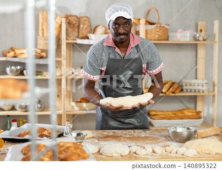 Professional male baker prepares raw yeast dough in bakery, shaping it into baguette Professional male baker prepares raw yeast dough in bakery, shaping it into baguette 140895322