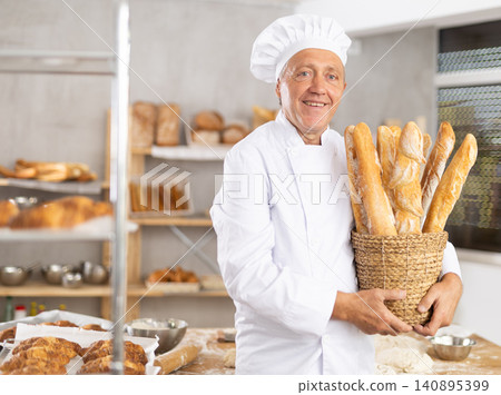 Smiling elderly baker presenting wicker basket of fresh baguettes Smiling elderly baker presenting wicker basket of fresh baguettes 140895399