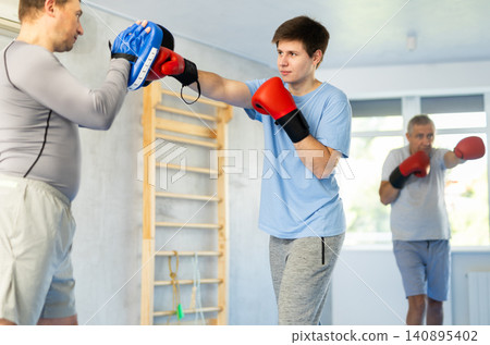 Guy in boxing gloves punching pads in hands of coach during workout Guy in boxing gloves punching pads in hands of coach during workout 140895402