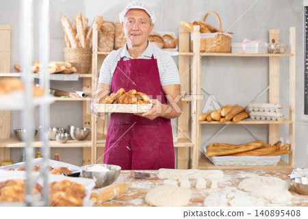 Smiling elderly baker in uniform stands behind counter in small private bakery offering croissants Smiling elderly baker in uniform stands behind counter in small private bakery offering croissants 140895408