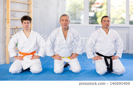 Sports team of different ages in white kimonos sit on their knees on the tatami in gym Sports team of different ages in white kimonos sit on their knees on the tatami in gym 140895409
