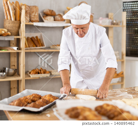 Professional older baker rolling dough on floured table Professional older baker rolling dough on floured table 140895462