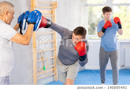 Man in boxing gloves punching mitts in hands of elderly instructor at gym Man in boxing gloves punching mitts in hands of elderly instructor at gym 140895491