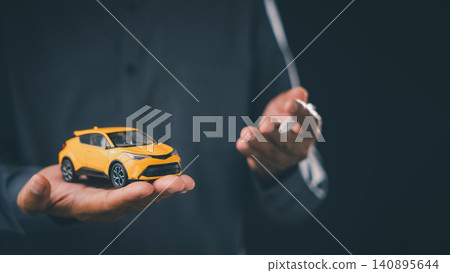 Dealership auto finance agent offers car buying loan approval and vehicle lease contract with insurance options while holding yellow toy car and pen against dark background 140895644