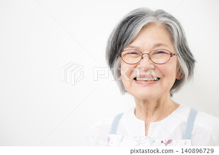 A close-up of a senior woman with gray hair, wearing an apron and a white background, smiling directly at the camera. A close-up of a senior woman with gray hair, wearing an apron and a white background, smiling directly at the camera. 140896729