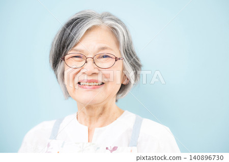 一位頭髮花白的老婦人,穿著圍裙,背景是淺藍色,對著鏡頭微笑,鏡頭特寫。 一位頭髮花白的老婦人,穿著圍裙,背景是淺藍色,對著鏡頭微笑,鏡頭特寫。 140896730