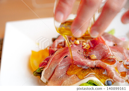 A hand pouring olive oil onto a prosciutto salad. A hand pouring olive oil onto a prosciutto salad. 140899161