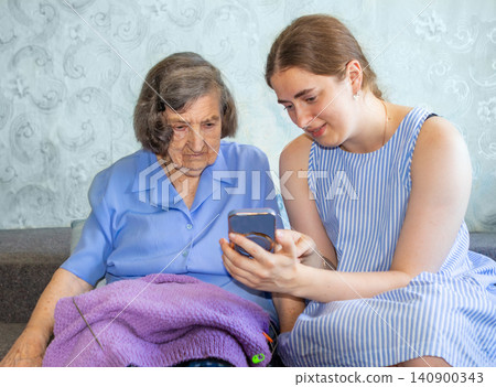 Granddaughter helps grandmother use smartphone while sitting together at home and sharing family care Granddaughter helps grandmother use smartphone while sitting together at home and sharing family care 140900343