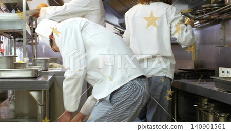 Reaching chef in white coat and striped trousers grabbing pan from lower shelf at busy kitchen Reaching chef in white coat and striped trousers grabbing pan from lower shelf at busy kitchen 140901561