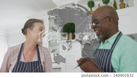 Talking senior man and mature woman smiling in home kitchen, with striped aprons and globe overlay Talking senior man and mature woman smiling in home kitchen, with striped aprons and globe overlay 140901632