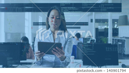 Standing woman wearing blue shirt checking tablet in open office, with monitors and code overlay Standing woman wearing blue shirt checking tablet in open office, with monitors and code overlay 140901794