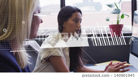 Pointing dark-haired woman in light top showing monitor overlay at office desk, peer in dark blazer Pointing dark-haired woman in light top showing monitor overlay at office desk, peer in dark blazer 140901945