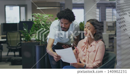 Reviewing colleagues holding paper and tablet at office near planters, white polo and pink blouse Reviewing colleagues holding paper and tablet at office near planters, white polo and pink blouse 140902231