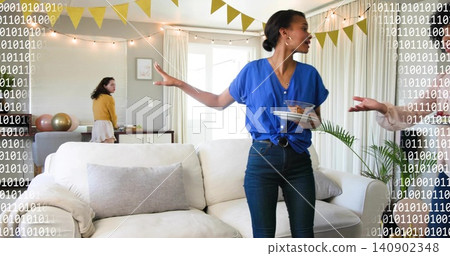 Gesturing woman in bright blue blouse holding plates and bowl at home living room, with bunting Gesturing woman in bright blue blouse holding plates and bowl at home living room, with bunting 140902348