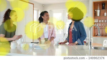 Holding white teapot, three adult women chatting and sipping at home kitchen island, wearing denim Holding white teapot, three adult women chatting and sipping at home kitchen island, wearing denim 140902374