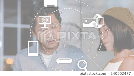 Working Asian colleagues in blue button-up and beige beanie examining AI icons in softly lit office 140902477