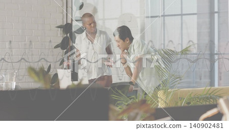 Discussing coworkers in light short sleeve tops at desk in open office with laptop, glass partition Discussing coworkers in light short sleeve tops at desk in open office with laptop, glass partition 140902481