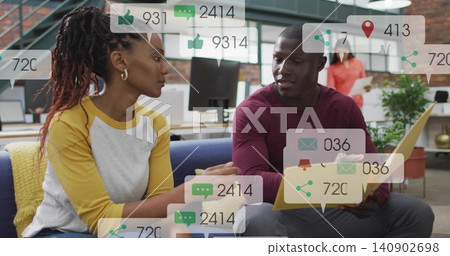 Discussing pair wearing maroon and yellow on blue sofa in office, using yellow laptop with overlays Discussing pair wearing maroon and yellow on blue sofa in office, using yellow laptop with overlays 140902698