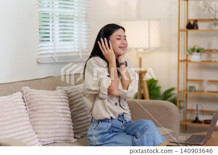 Asian woman relaxing on sofa listening to music with headphones at home Asian woman relaxing on sofa listening to music with headphones at home 140903646