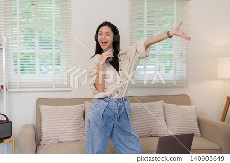 Asian woman wearing headphones singing and dancing in living room for leisure Asian woman wearing headphones singing and dancing in living room for leisure 140903649