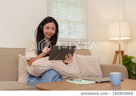Asian woman wearing headphones using digital tablet on sofa for remote work 140903669