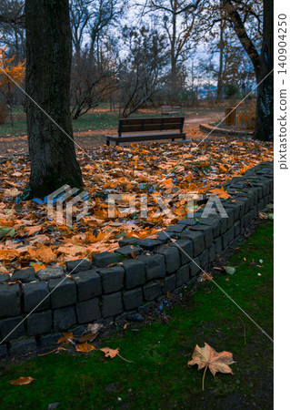 Autumn park square with fallen leaves and empty bench under cloudy skies 140904250