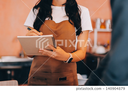 Friendly Asian female barista receives a credit card from a customer for payment. Both women are smiling in a warmly lit coffee shop with a rustic interior. Cashless payments. Small family business Friendly Asian female barista receives a credit card from a customer for payment. Both women are smiling in a warmly lit coffee shop with a rustic interior. Cashless payments. Small family business 140904879