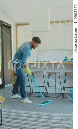 Vertical Video Man in protective gloves is singing using a mop and dancing while cleaning his house in living room during end of year clean. Vertical Video Man in protective gloves is singing using a mop and dancing while cleaning his house in living room during end of year clean. 140905353