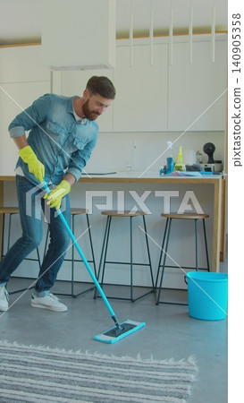 Vertical Video Man in protective gloves is singing using a mop and dancing while cleaning his house in living room during end of year clean. Vertical Video Man in protective gloves is singing using a mop and dancing while cleaning his house in living room during end of year clean. 140905358