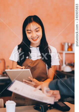 Friendly Asian female barista receives a credit card from a customer for payment. Both women are smiling in a warmly lit coffee shop with a rustic interior. Cashless payments. Small family business Friendly Asian female barista receives a credit card from a customer for payment. Both women are smiling in a warmly lit coffee shop with a rustic interior. Cashless payments. Small family business 140905663