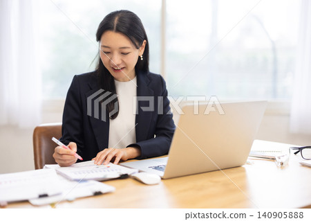 A businesswoman writing notes on documents with a pen while working on her computer. A businesswoman writing notes on documents with a pen while working on her computer. 140905888