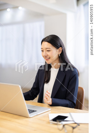 A young Japanese businesswoman smiles and talks while looking at her computer by a bright window. A young Japanese businesswoman smiles and talks while looking at her computer by a bright window. 140905955