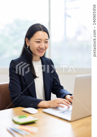 A young Japanese businesswoman smiles and talks while looking at her computer by a bright window. A young Japanese businesswoman smiles and talks while looking at her computer by a bright window. 140905956