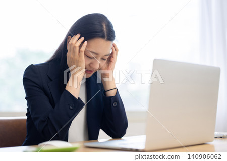 一位年輕的日本女商人坐在明亮的窗邊辦公桌前,雙手托著太陽穴,低頭看著眼前的電腦。 一位年輕的日本女商人坐在明亮的窗邊辦公桌前,雙手托著太陽穴,低頭看著眼前的電腦。 140906062