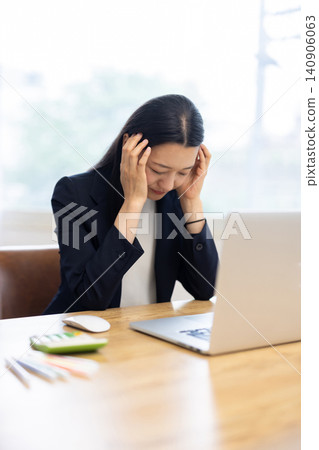 一位年輕的日本女商人坐在明亮的窗邊辦公桌前，雙手托著太陽穴，低頭看著眼前的電腦。 140906063