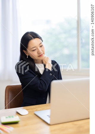 A young Japanese businesswoman, working on her laptop by a bright window, touches her shoulder, appearing to be in pain. 140906107