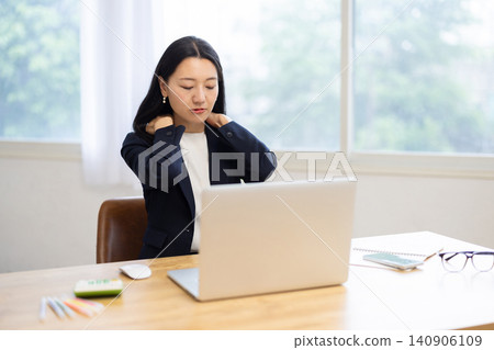 A young Japanese businesswoman, working on her laptop by a bright window, touches her shoulder, appearing to be in pain. A young Japanese businesswoman, working on her laptop by a bright window, touches her shoulder, appearing to be in pain. 140906109
