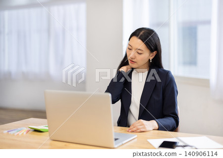 一位年輕的日本女商人坐在明亮的窗邊用筆記型電腦工作，她摸了摸自己的肩膀，似乎很痛苦。 140906114