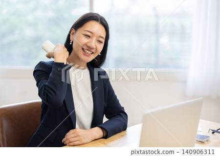 一位年輕的日本女商人坐在辦公桌旁,靠著明亮的窗戶,用按摩槍為自己按摩。 一位年輕的日本女商人坐在辦公桌旁,靠著明亮的窗戶,用按摩槍為自己按摩。 140906311