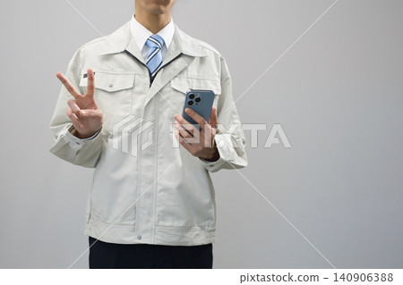 一名身穿工作服的年輕男子手持智慧型手機,比出V字手勢。 一名身穿工作服的年輕男子手持智慧型手機,比出V字手勢。 140906388