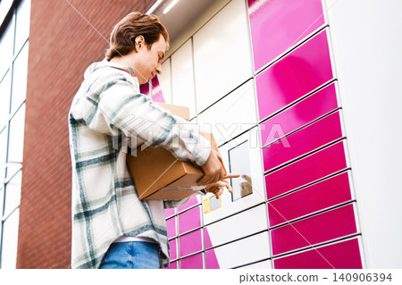 man holding a box uses a screen at a parcel locker for his delivery pickup. This automated terminal makes ecommerce logistics and contactless online shopping easy. man holding a box uses a screen at a parcel locker for his delivery pickup. This automated terminal makes ecommerce logistics and contactless online shopping easy. 140906394