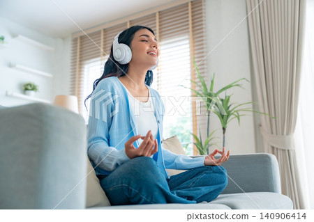 Happy young Asian woman practicing yoga and meditation at home sitting on couch in living room Happy young Asian woman practicing yoga and meditation at home sitting on couch in living room 140906414