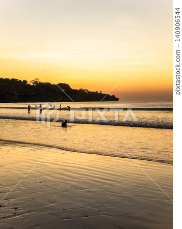 People Swimming in Ocean Waves at Tropical Beach During Vivid Orange Sunset with Tree Silhouette 140906544