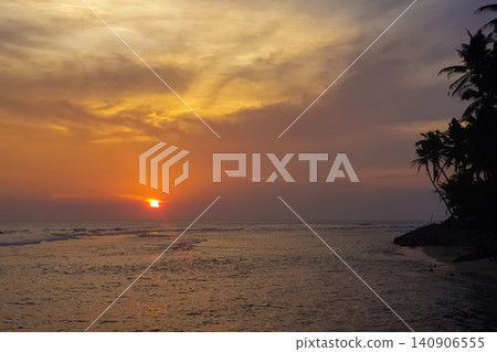 Tropical beach sunset with palm tree silhouettes, orange sky and calm ocean waves on a shoreline Tropical beach sunset with palm tree silhouettes, orange sky and calm ocean waves on a shoreline 140906555